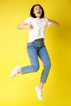 Full size of happy young asian woman jumping from joy, showing thumbs up in approval, posing over yellow background in jeans and casual white t-shirt.