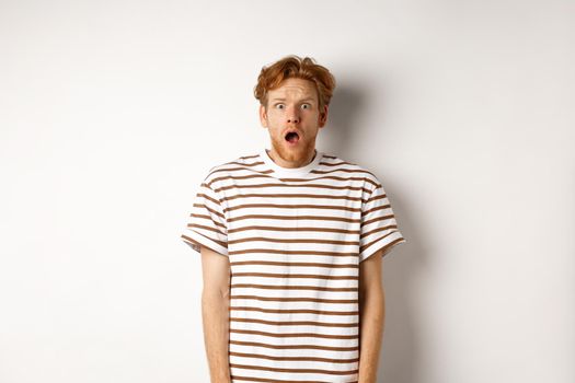 Shocked and scared redhead man screaming, open mouth and staring terrified at camera, standing over white background.