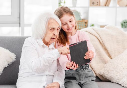 Grandmother with grandaughter sitting on the sofa, looking at tablet screen and smiling