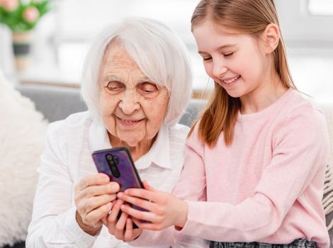 Grandmother with grandaughter sitting together, looking at smartphone and smiling