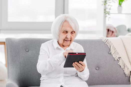 Portrait of elderly gey hair woman sitting on the sofa at home with tablet and looking at screen