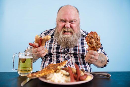Angry mature bearded man with overweight holds smoked chicken leg and sausages at table with greasy food on light blue background in studio