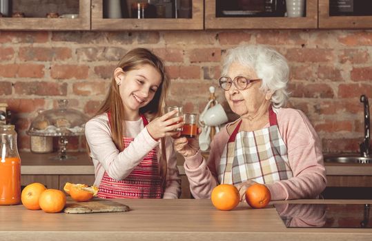 Granny with happy granddaughter tasting and drinking fresh natural orange juice at kitchen at home