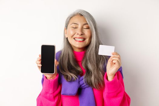 Online shopping. Happy and delighted asian senior woman smiling with joy, showing blank smartphone screen and plastic card, buying in internet, white background.