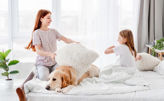 Two sisters fighting with pillow in the bed and golden retriever dog lying close to them. Girls have a fun in the morning in sunny room with daylight and pet doggy resting