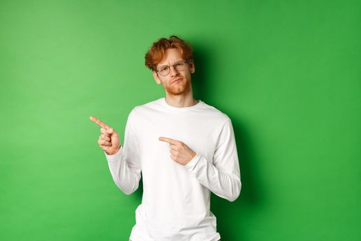 Doubtful redhead guy in glasses pointing fingers left, looking hesitant and unsure at camera, standing over green background.