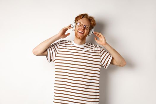 Technology concept. Happy redhead man listening music in headphones and singing along, standing over white background.