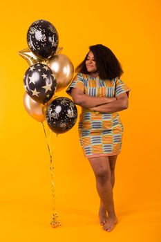 Portrait of smiling young African-American adult woman looking sweet on yellow background holding balloons.