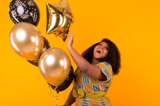 Portrait of smiling young African-American adult woman looking sweet on yellow background holding balloons.