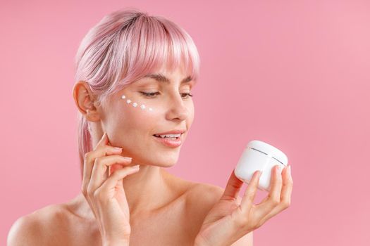 Portrait of beautiful woman with pink hair and face cream applied on her skin like dots holding and looking at cream in white jar isolated over pink background. Beauty, spa, skin care concept