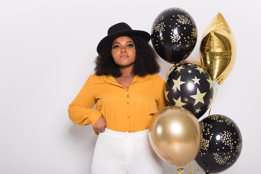 Portrait of smiling young African-American adult woman looking sweet on yellow background holding balloons.