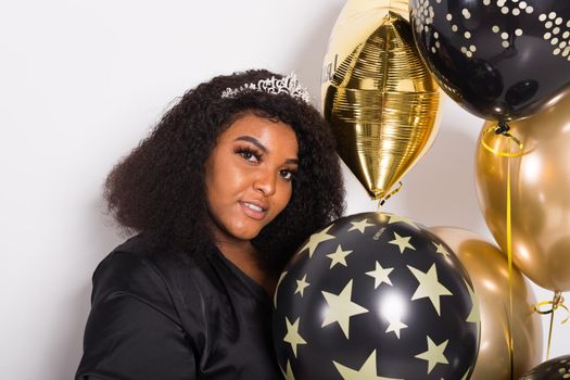 Portrait of smiling young African-American adult woman looking sweet on yellow background holding balloons.