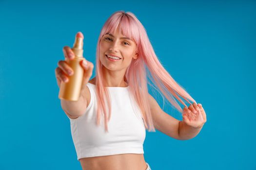 Portrait of young woman with pink hair smiling and showing bottle of moisturizing spray to camera while standing isolated over blue studio background. Beauty products, hair care concept