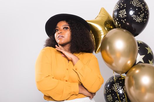 Portrait of smiling young African-American adult woman looking sweet on yellow background holding balloons.