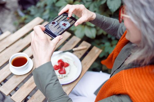 Senior woman guest takes picture of beautiful strawberry dessert with smartphone sitting at table on outdoors cafe terrace on autumn day