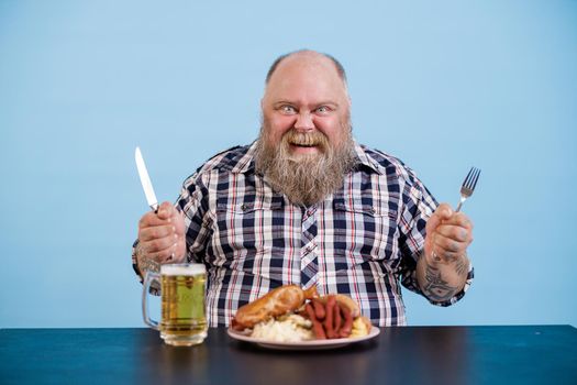 Joyful bearded person with overweight holds knife and fork sitting at table with rich food and glass of beer on light blue background in studio