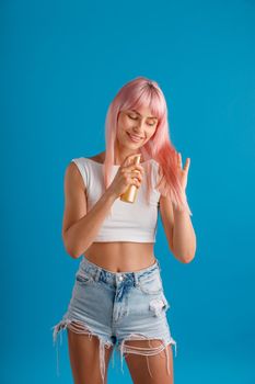 Cheerful young woman smiling and applying moisturizing spray to her beautiful smooth pink dyed hair while standing isolated over blue studio background. Beauty products, hair care concept