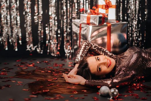 Top view of a girl lying in shiny clothes on the floor in confetti in the form of hearts and gifts.