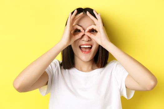 Close up of funny asian girl looking through hand binoculars and smiling, standing in t-shirt against yellow background.