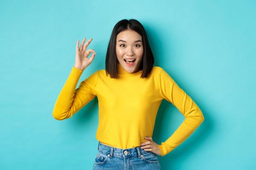 Amazed korean girl showing okay gesture and smiling at camera, approve and recommend special deal, standing over blue background.