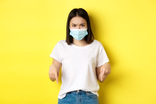 Covid-19, pandemic and social distancing concept. Disappointed asian girl in medical mask, frowning upset and pointing fingers down at logo, standing over yellow background.