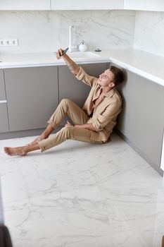 Beautiful caucasian man is sitting on the floor and using smartphone on light modern kitchen