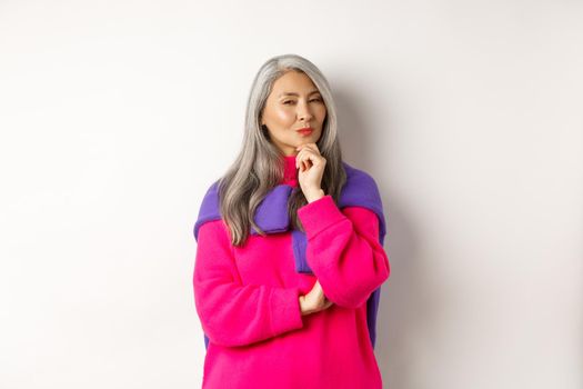 Image of thoughtful asian senior woman squinting suspicious at camera, having assumption, standing over white background and thinking.