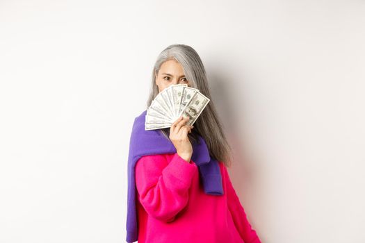 Shopping concept. Rich and fashionable korean senior woman hiding face behind money, looking at camera, standing over white background.