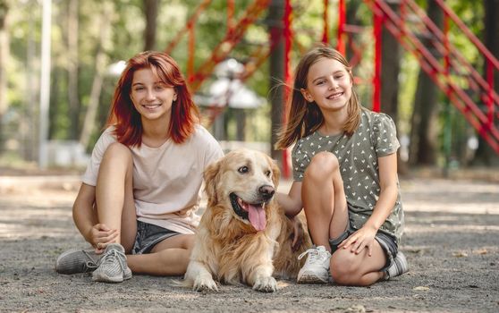 Beautiful girls with golden retriever dog sitting in the park and smiling. Sisters with doggy pet outdoors