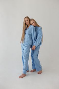 two young twin sisters with long blond hair posing on white background in oversize clothes with bare feet and looking at camera. they wear blue suits that look like pajamas and hug. fashion photoshoot