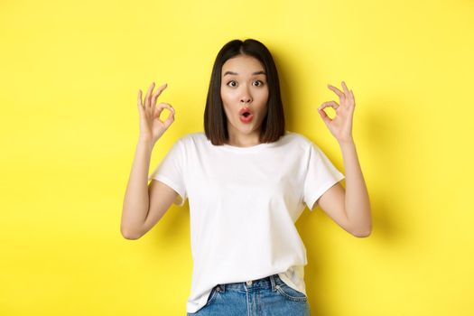 Impressed korean girl saying WOW, showing okay signs and looking amazed, standing against yellow background.