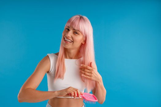 Cheerful woman brushing her smooth natural long pink hair with hair comb while posing isolated over blue studio background. Beauty, hair care concept