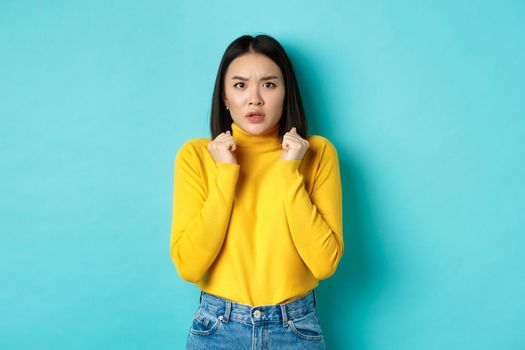 Image of worried asian woman with short dark hair, clench hands and stare at camera concerned, standing over blue background.
