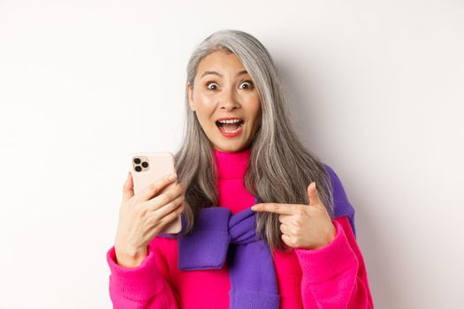 Online shopping. Close up of excited asian senior woman pointing at smartphone and smiling amazed, standing over white background.