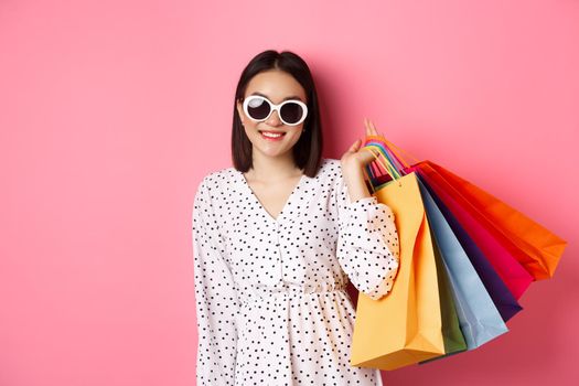 Young asian woman in sunglasses going shopping, holding bags from malls and stores and smiling, standing over pink background.