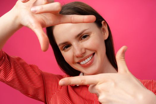 Smiling young woman showing frame with fingers against pink background, close up