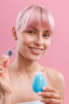 Portrait of happy young woman with pink hair and perfect skin holding dropper and bottle of serum or hyaluronic acid, posing isolated over pink background. Beauty, cosmetics and skincare concept