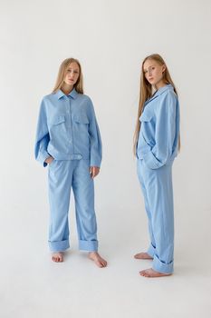 two young twin sisters with long blond hair posing on white background in oversize clothes with bare feet and looking at camera. They wear blue suits that look like pajamas. Stylish fashion photoshoot