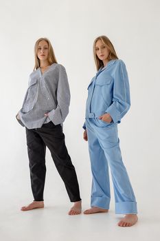 two cute young twin sisters with long blond hair posing on white background in oversize clothes with bare feet. Blue suit on one girl, striped shirt and black pants on another one. Fashion photoshoot