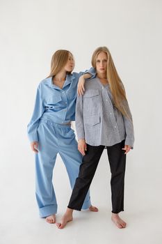 two cute young twin sisters with long blond hair posing on white background in oversize clothes with bare feet. Blue suit on one girl, striped shirt and black pants on another one. Fashion photoshoot