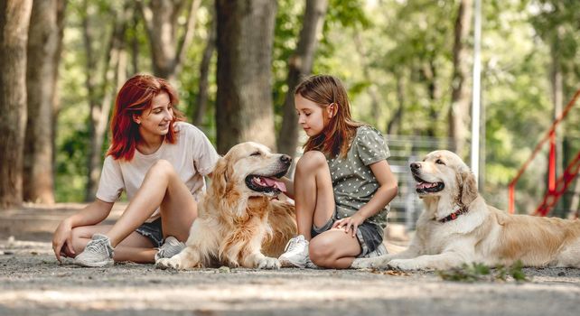 Girls sisters with golden retriever dogs sitting in the park. Family with doggy pets outdoors