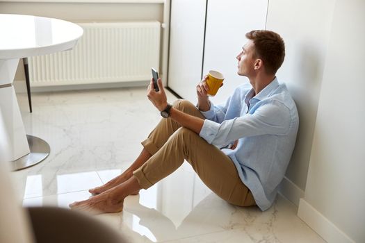 Caucasian hadsome young man in blue shirt is sitting on the floor and chilling at modern kitchen