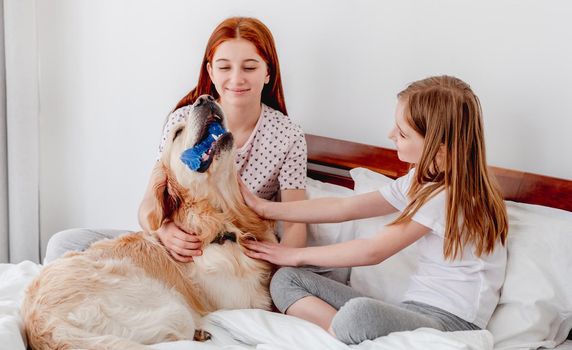 Two sisters sitting in the bed and petting golden retriever dog while doggy holding in his teeth toy bone. Girls with pet staying at home. Beautiful portrait of friendship between human and animal