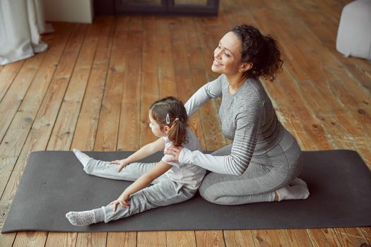 Top view of happy mother helping sporty little girl during domestic gymnastics training on mat