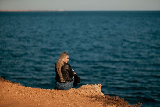 A blonde girl in a stylish black leather jacket is sitting with her back to the seashore