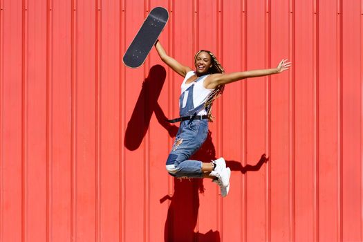 Black young woman wtih a skateboard jumping with happiness on red urban wall background.