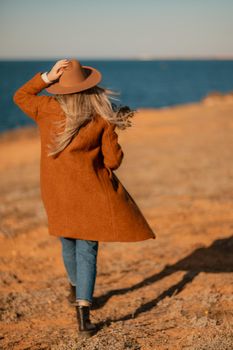 A woman walking along the coast near the sea. An elegant lady in a brown coat and a hat with fashionable makeup walks on the seashore.