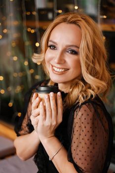 Young charming blonde with a cute smile and makeup while relaxing in a cafe. She is holding a cup of coffee in her hands. She is dressed in a black dress with transparent sleeves