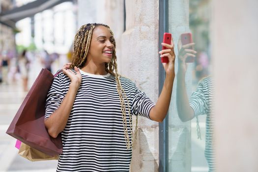 Young black woman photographing a store window in a shopping street with her smartphone. Female with african braids.