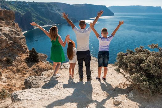 Family mom dad and two children, a boy and a girl stand with their backs to the audience and look out over the sea. They raised their hands. Against the backdrop of a beautiful seashore with rocks